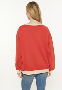 Yli suuri punainen huppari, jossa on leveä kaula-aukko, tilavat hihat ja vaalean kermaoinen ribbireuna. Pehmeä kangas; rento muotoilu.