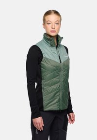 Gilet verde imbottito con motivo a zigzag sul frontale, colletto alto e tasche con zip. Tessuto leggero con un sottostrato nero a contrasto.