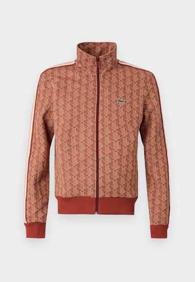 Veste zippée à motifs rouges avec un col haut, des poignets et un ourlet côtelés. Présente des rayures latérales contrastantes et un petit logo sur la poitrine.