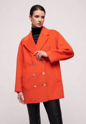 Orange wool blend coat with a double-breasted design, large lapels, two pockets, and gold-tone buttons. Model wearing a black striped top.