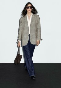 Blazer a scacchi grigio sopra una camicia bianca, abbinato a pantaloni ampi in denim scuro. Borsa a spalla marrone, scarpe nere, capelli lunghi e mossi, occhiali da sole scuri.