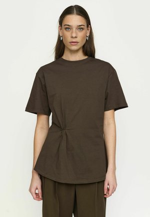 Žena s dlhými hnedými vlasmi, ktorá nosí strieborné kruhové náušnice, tmavohnedé tričko s krátkym rukávom so zaväzovaním na boku a zodpovedajúce nohavice.