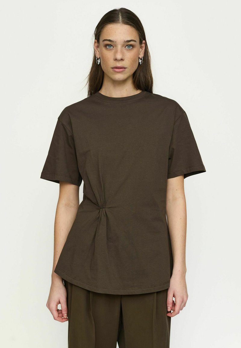 Femme aux longs cheveux bruns portant des boucles d'oreilles argentées en anneau, un t-shirt marron foncé à manches courtes avec un nœud latéral, et un pantalon assorti.