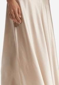 En golvlång, ljusbeige satin kjol med en slät textur, som har subtila plisseringar och en hand som försiktigt vilar på dess yta.