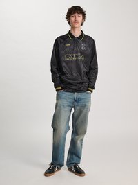 Ung mann står med hendene i lommene, iført svart sportstrøye med lange ermer, falmet blå jeans og svarte joggesko med hvite striper.