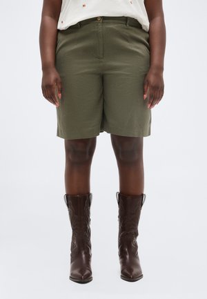 Personne portant un short mi-long de couleur vert olive associé à des bottes de cowboy marron foncé à motifs, debout devant un fond uni.