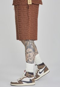Braun gemusterte Shorts mit weißen und grauen Nike Sneakern. Ein detailliertes Porträt-Tattoo am Bein. Weiße, gerippte Socken getragen.