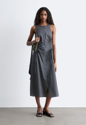 Mujer con pelo largo y rizado que lleva un vestido midi sin mangas a rayas con botones, zapatos de tacón abiertos negros y lleva dos bolsos de hombro.