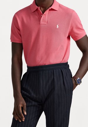 Personne à la peau foncée portant un polo rose à manches courtes avec un logo blanc, un pantalon foncé à fines rayures et une montre-bracelet au cadran bleu avec un bracelet marron.