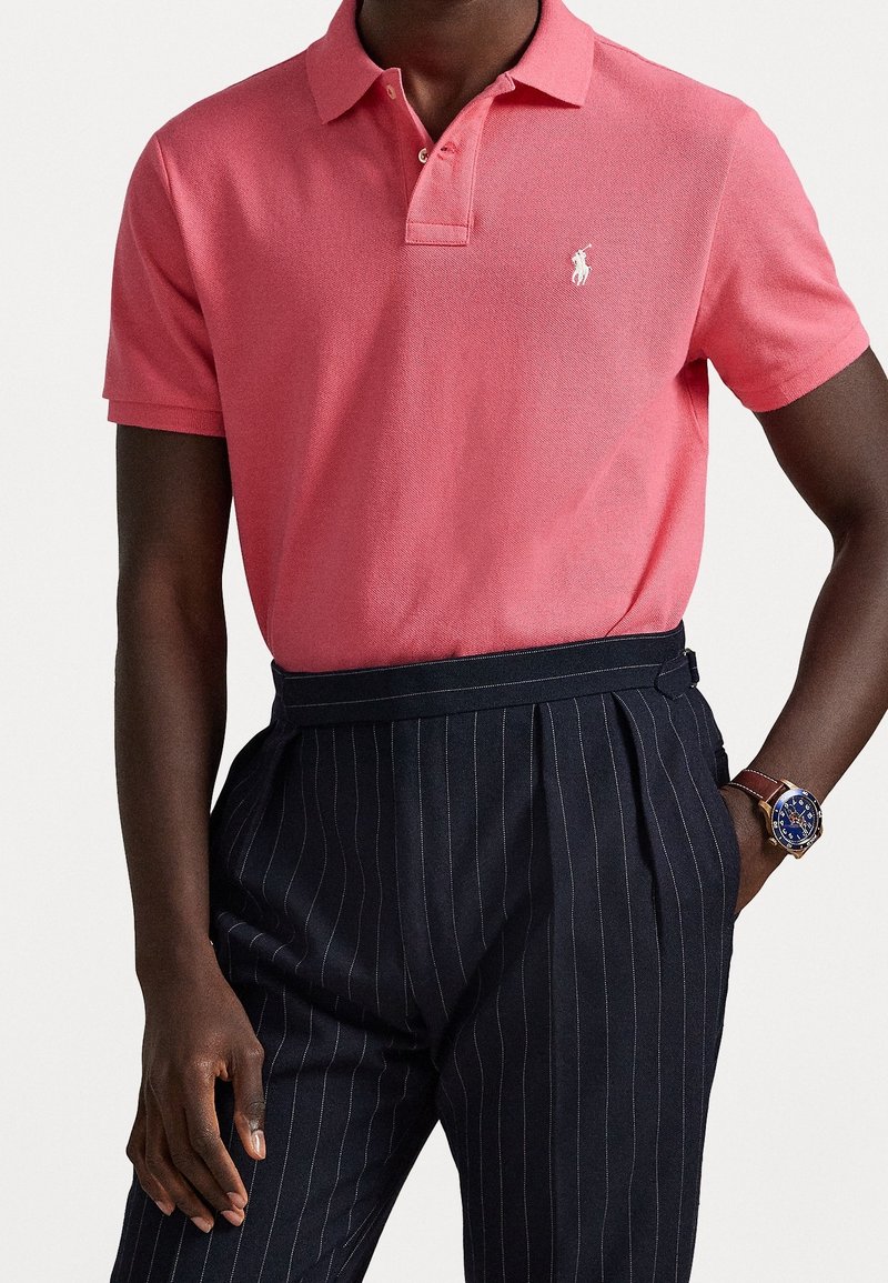 Personne à la peau foncée portant un polo rose à manches courtes avec un logo blanc, un pantalon foncé à fines rayures et une montre-bracelet au cadran bleu avec un bracelet marron.