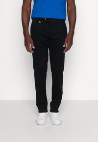 Pantaloni neri in cotone con taglio slim, dotati di zip e chiusura con bottone, indossati con una camicia blu e sneakers bianche.