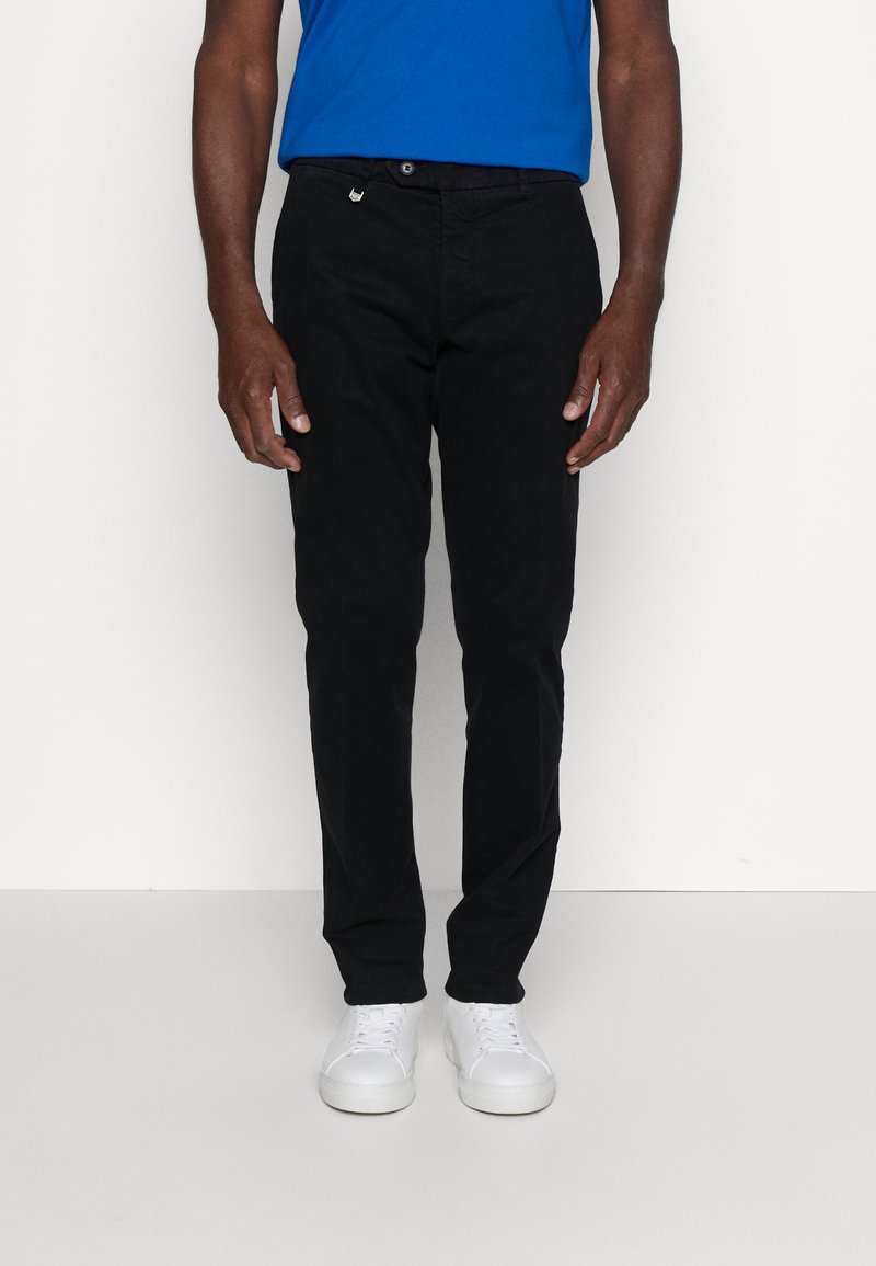Pantaloni neri in cotone con taglio slim, dotati di zip e chiusura con bottone, indossati con una camicia blu e sneakers bianche.