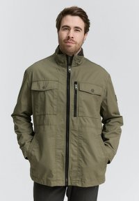 Olivgrüne Jacke mit einem hohen Kragen, Frontreißverschluss, mehreren Taschen und einer lässigen Passform. Der Stoff wirkt glatt mit minimalen Texturdetails.