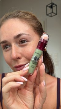 Junge Frau hält einen grünen Maybelline Farbkorrektur-Concealer mit Schwammapplikator in der Nähe ihres Gesichts, zeigt natürliche Haut und gepflegte Nägel.