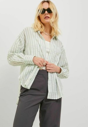 Camisa de botones de rayas verde claro y blanca con mangas largas, combinada con pantalones oscuros grises. La camisa tiene un corte relajado y un bolsillo.