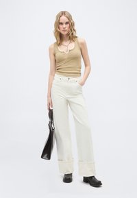 Femme portant un débardeur beige et un pantalon blanc à jambes larges avec des revers, tenant un sac à main noir et portant des bottines noires, se tenant devant un fond blanc.