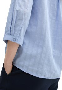 Light blue, striped blouse with three-quarter sleeves, buttoned cuffs, and soft texture. Worn with dark pants, hand in pocket.