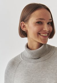 Maglione a collo alto grigio con colletto a coste, texture liscia e dettagli di cucitura sottili. Il modello ha i capelli corti e sorride.