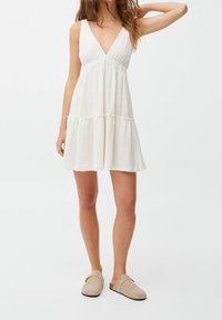 Robe blanche sans manches avec un décolleté en V, un design à étages et un tissu texturé. Portée avec des chaussures beiges à enfiler.
