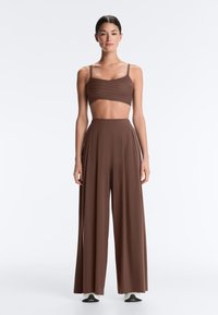 Femme aux cheveux foncés portant un débardeur court marron et un pantalon marron taille haute à jambes larges, debout sur un fond blanc uni.