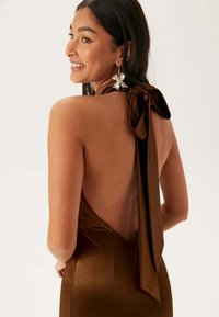 Brun satin klänning med öppen rygg och en stor rosett vid halsen. Har en slät struktur och figurnära design. Utmärkande blommiga örhängen.