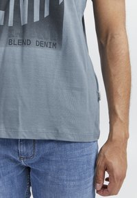 Grå T-skjorte med grafisk trykk i mørkere nyanser, laget av myk bomull, med korte ermer og hempeetikett. Brukt med blå denimjeans.