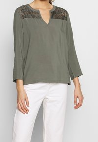 Top verde oliva con scollatura a V e maniche lunghe, caratterizzato da dettagli in pizzo nero intricati sulle spalle, tessuto morbido e vestibilità comoda.