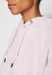 Lyserosa fleece hettegenser med myk tekstur, med runde snorer med svarte tips og en merketag på halsen.