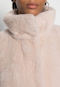 Abrigo de pelo sintético en rosa claro con un alto cuello. Presenta una textura suave y una costura central, complementado por un acabado liso.