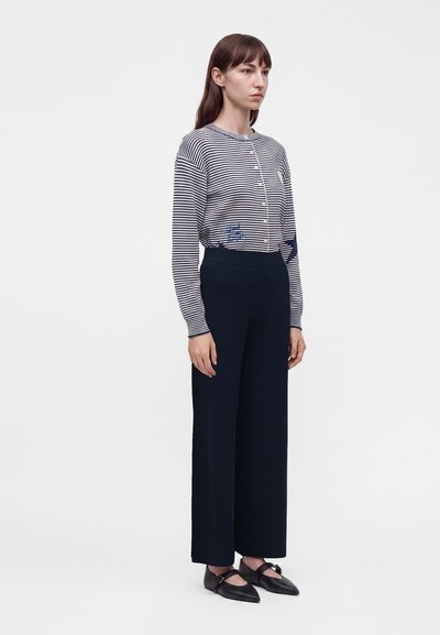 Femme debout portant un haut à manches longues rayé et boutonné, un pantalon noir taille haute à jambes larges, et des chaussures Mary Jane noires pointues.