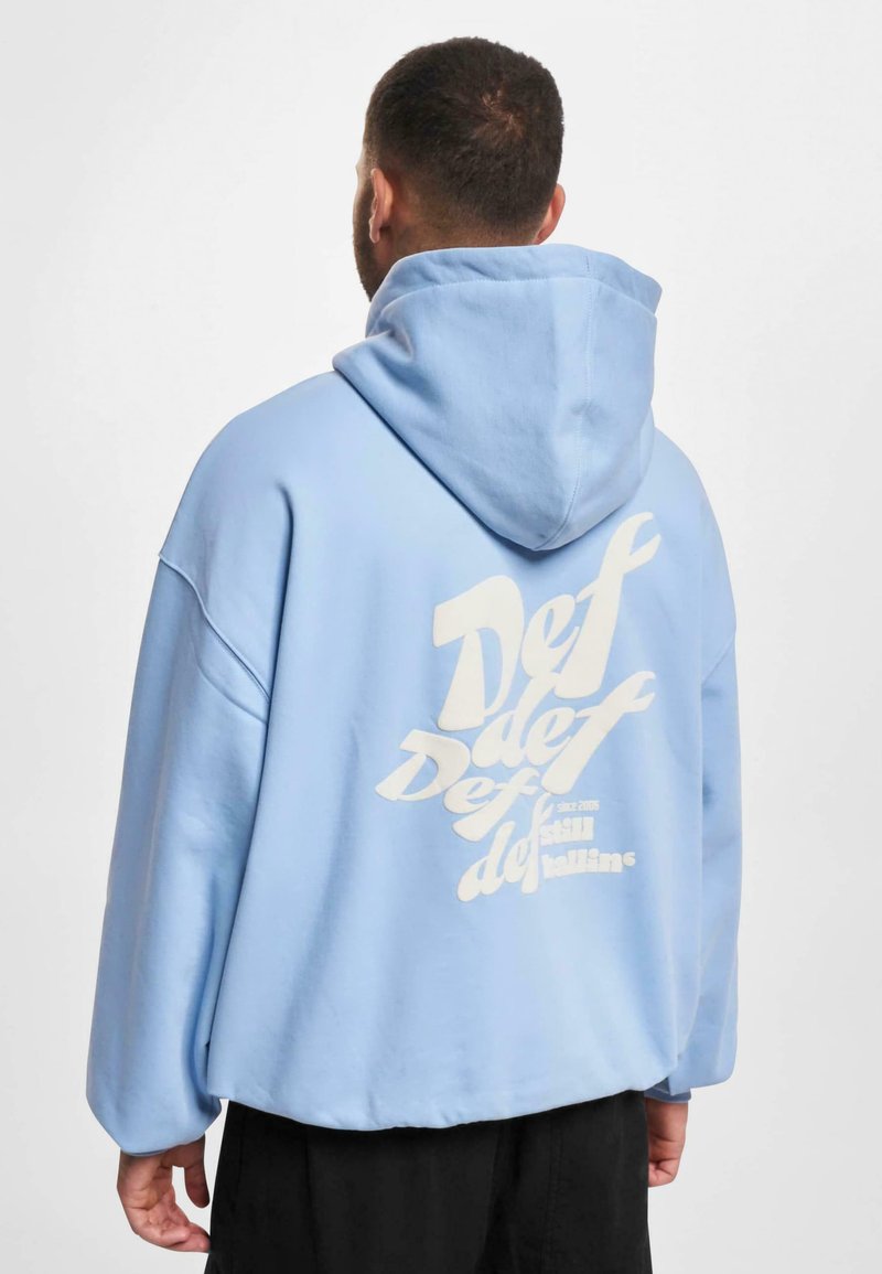 Lys blå oversized hettegenser med en frontlomme og hvit grafisk tekst på baksiden. Stoffet ser mykt og glatt ut.