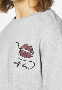 Sudadera gris con un diseño de labios de lentejuelas en tonos rosa y negro, con bordado azul que dice "amor propio" debajo de la gráfica.