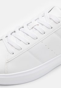 Hvide lædersneakers med flade hvide snørebånd, syet detaljer og subtile perforeringer på siden. Gummisål med tekstureret greb.