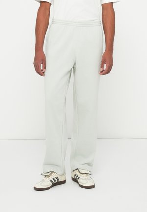 Man in lichtgrijze sweatpants en off-white sneakers met zwarte strepen, staand tegen een eenvoudige witte achtergrond.