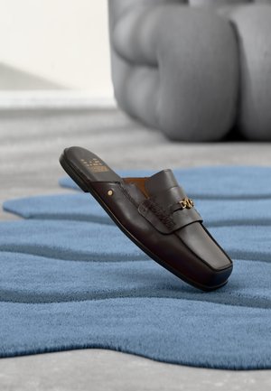 Mocassin à enfiler en cuir marron foncé avec détail décoratif en chaîne dorée sur une surface texturée bleue, sculpture moderne floue en arrière-plan.