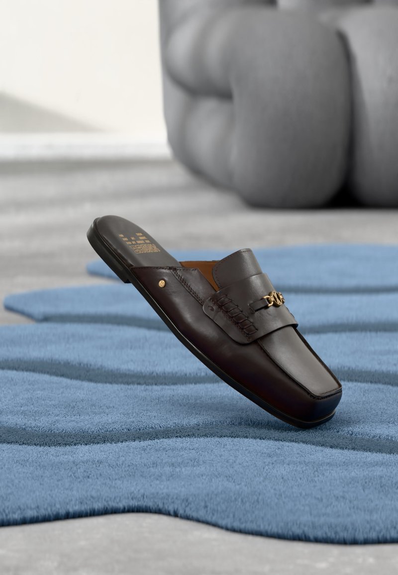 Mocassin à enfiler en cuir marron foncé avec détail décoratif en chaîne dorée sur une surface texturée bleue, sculpture moderne floue en arrière-plan.