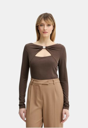 Donna con capelli biondi lisci che indossa una maglia marrone a maniche lunghe con un ritaglio frontale e pantaloni beige a vita alta su uno sfondo semplice.