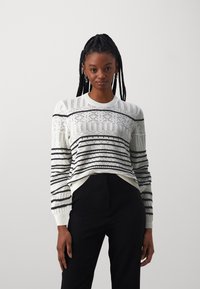 White knitted sweater with black stripes and geometric patterns, featuring a round neckline and long sleeves, worn over black pants.