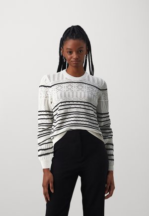 Pulover alb tricotat cu dungi negre și modele geometrice, având guler rotund și mâneci lungi, purtat peste pantaloni negri.