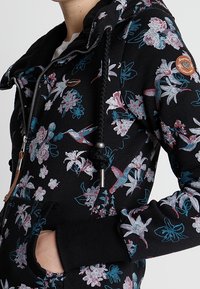 Zwarte hoodie met bloemendessin in roze en blauw. Voorzien van een rits, trekkoordkap en frontzakken. Textuurstof.