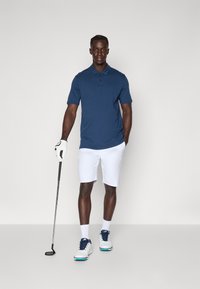 Golfový outfit obsahující námořnickou polokošili, bílé kraťasy, bílé golfové boty s modrými akcenty a golfovou rukavici. Postoj ve stoje s golfovou holí.