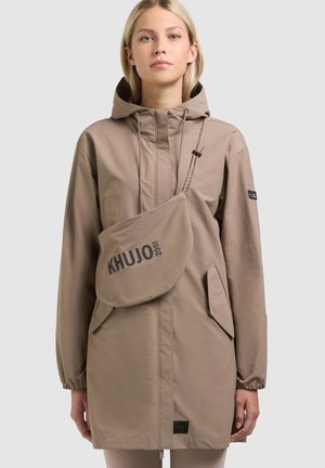 Junge Frau trägt eine taupefarbene Kapuzenjacke und eine passende Umhängetasche mit der Aufschrift "KHUJO 2001", stehend vor einem grauen Hintergrund.