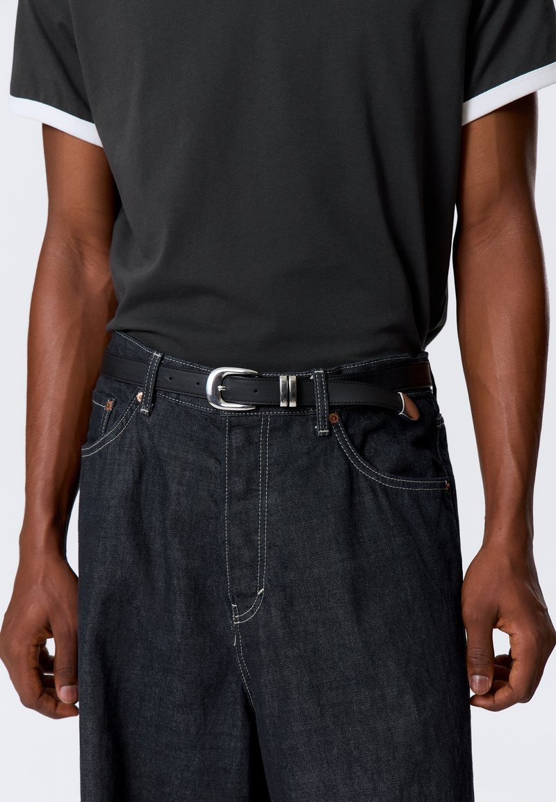 Personne portant un t-shirt noir rentré dans un jean en denim foncé avec une ceinture en cuir noire et une boucle argentée.