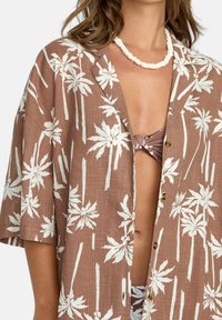Camicia in lino marrone con stampa di palme bianche, maniche corte e vestibilità rilassata. Bottoni visibili lungo la parte frontale per la chiusura.