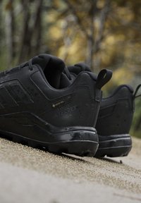Coppia di scarpe da trekking nere Gore-Tex Terrex su una superficie ghiaiosa con alberi autunnali sfocati sullo sfondo.