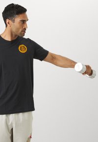 Schwarzes T-Shirt mit kurzen Ärmeln und orangefarbenem Logo, getragen von einer Person, die eine silberne Hantel hebt, vor einem neutralen Hintergrund.