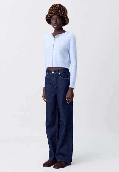 Maglione accorciato azzurro chiaro con chiusura a bottoni, abbinato a jeans in denim scuri a gamba larga. Il modello indossa un cappello a secchiello in stampa leopardo marrone.