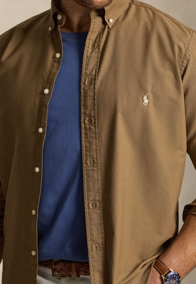 Mężczyzna w rozpiętej brązowej koszuli na niebieskim t-shircie, z plecionym brązowym paskiem i zegarkiem z niebieską tarczą na lewej ręce.