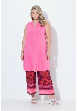 Kvinde iført ærmeløs lyserød knap-tunika, viddebukser med blomsterprint i rød og pink, og hvide sandalettes med hæl, stående mod hvid baggrund.