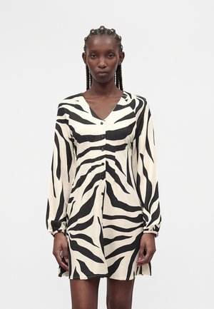 Femme aux cheveux tressés portant une robe boutonnée à manches longues, noire et blanche à rayures zébrées, debout devant un fond clair uni.